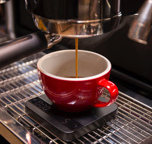Extracción de espresso con porta naked (desnudo) en taza roja con báscula para medir los gramos de café líquido de acuerdo con la fórmula o receta para el espresso. Curso para baristas de Kima Coffee en Málaga